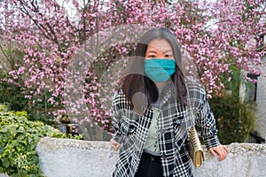 Traveling in virus times - young happy and beautiful Asian Korean woman in face mask against covid19 enjoying sakura blooming tree