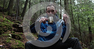 Traveling young tourist enjoying nature drinking hot tea while sitting in forest in rainy