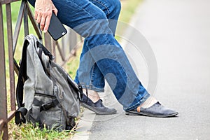 Traveller legs in jeans and hand with cellphone, man resting and sitting steel fence, backpack laying on ground