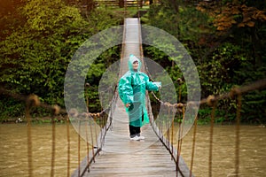 Traveller senior beautiful woman in blue rain jacket cross river by hinged bridge in forest, enjoying nature