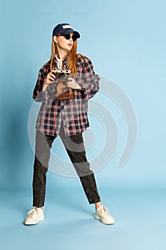 Traveller. Young beautiful girl in checkered shirt, cap and sunglasses posing with photo camera over blue studio