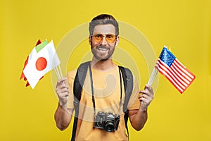 Travelling abroad. Male tourist holding international flags, standing with backpack and camera over yellow background