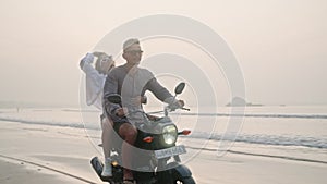 Travelling couple rides motorcycle on wet sand sea beach at sunrise. Man, woman drive rented scooter on coastline along