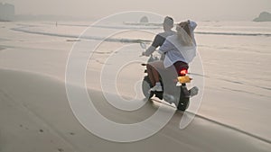 Travelling couple rides motorcycle on wet sand sea beach at sunrise. Man, woman drive rented scooter on coastline along