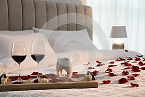 Tray with glasses of red wine, candles and rose petals on bed in room