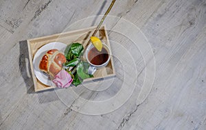 Tray with lemon tea and hot cross bun with a pink rose lying across the try. Set on a grey wooden background. A treat for easter