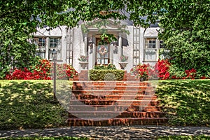 Tree shaded dappled sidewalk and sloped lawn in front of upscale stucco house with wreath and azaleas and brick steps - beautiful