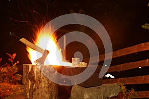 Tree stump with axe and burning firewood in metal brazier outdoors at night
