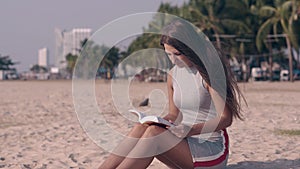 Tremendous girl in light dress reads book sitting on sand