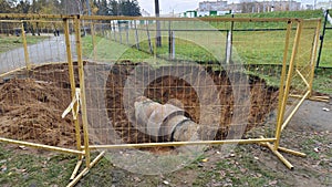 A trench for replacing sewer pipes and other utilities has been dug in a lawn with fallen fall leaves and a dirt path and fenced o