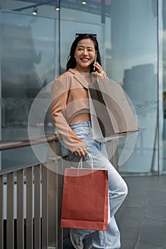 A trendy Asian woman is talking on the phone with her friend during her shopping day at the mall