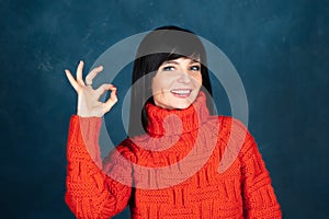 Trendy beautiful girl, brunette in a bright orange sweater, shows an ok sign and smiling . The theme of autumn and coolness.