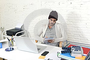 Trendy businessman in cool hipster beanie writing on pad working in at modern home office with computer