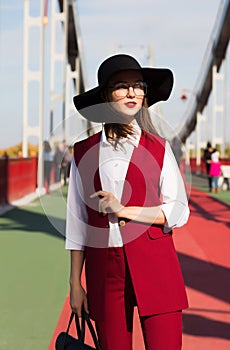 Trendy dressed brunette girl in red pantsuit and black hat posing at the bridge