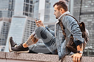 Trendy dressed tattooed hipster man with a backpack using smartphone in front of skyscrapers in Moskow city at cloudy
