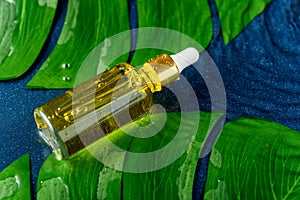 Trendy dropper bottle with white pipette and yellow liquid on classic blue background with water drops and green leaf