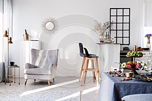 Trendy grey armchair next to two black wooden bar stools in fashionable kitchen and dining room