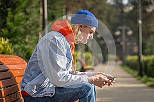 Trendy guy sitting on bench in park, waiting for girlfriend, surfing in mobile internet, messaging