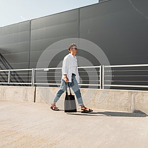 Trendy hipster young man in sunglasses in fashionable summer clothes with cloth bag in vintage sandals walks on the street