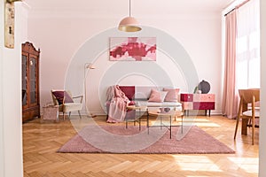 Trendy living room interior with grey couch with pastel pink pillows and blanket, stylish beige armchair with burgundy pillow and