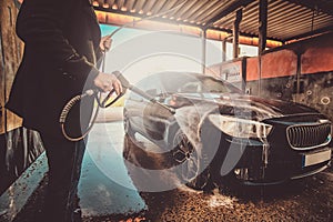 Trendy man in jeans and blaser is washing his own car at car washing station.