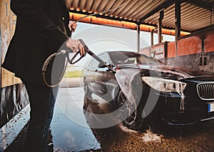 Trendy man in jeans and blaser is washing his own car at car washing station.