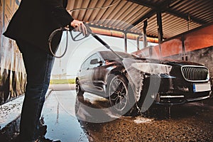 Trendy man in jeans and blaser is washing his own car at car washing station.