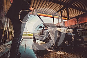 Trendy man in jeans and blaser is washing his own car at car washing station.