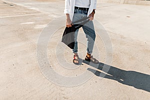 Trendy men`s legs in blue vintage jeans in stylish red leather sandals with a black fabric bag stand on the pavement. Fashionable