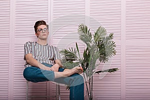 Trendy modern young man with a fashionable hairstyle in stylish youth clothes in vintage glasses is resting on a chair next to a