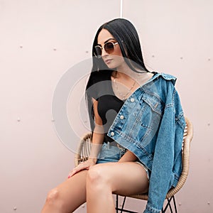Trendy pretty young hipster woman in stylish sunglasses in a fashionable denim jacket is sitting on a vintage straw chair