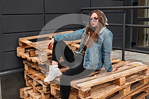 Trendy pretty young woman hipster in violet sunglasses in military hood in fashion denim jacket in jeans in white leather boots