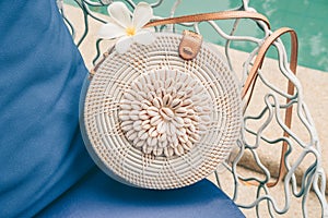 Trendy round crossbody straw bag with seashells and flower on the beautiful lounge chair near a pool