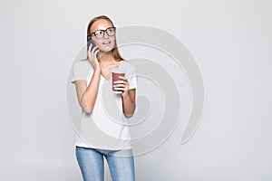 Trendy slender young woman standing chatting on a mobile phone while holding a mug of takeaway coffee in her other hand, isolated