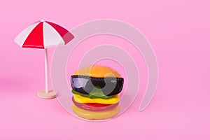 Trendy summer concept with fast food - hamburger in sunglasses and sun umbrella  on pastel pink background