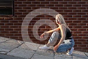 Trendy teen blonde girl in casual clothes crouched near the red brick wall, sun rays are falling on her face