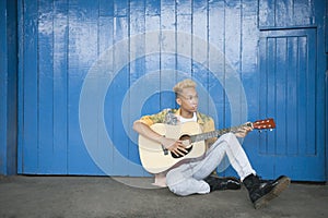 Trendy teenage boy playing guitar as he sits against wood paneled wall