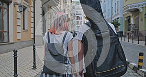 Trendy teenage hipsters boy and girl walking talking on city street, boy with guitar in case, back view