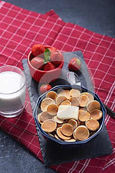 Trendy tiny cereal pancakes with slice of butter in blue bowl, strawberries, glass of milk on red background, rural Breakfast