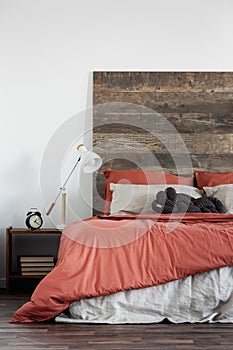 Trendy white lamp next to black clock on wooden nightstand next to king size bed with wooden headboard