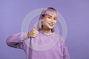 Trendy woman showing thumb up like sign over violet studio background. Positive young girl with dyed purple hair smiles