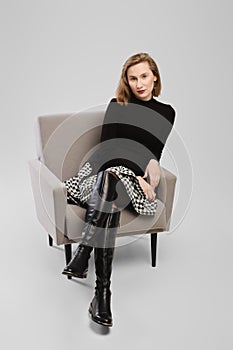 Trendy woman in turtleneck and ittle skirt sitting in armchair in studio and looking condescending
