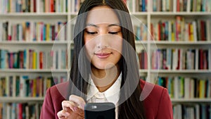 Trendy woman using her phone in a library to research. Young, stylish student browsing an online ebook reader app or