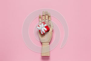 Trendy wooden mannequin hand holds small red gift box on pastel pink background, copy space, flat lay. March 8th, February 14th,