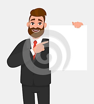 Trendy young business man showing blank white poster and pointing finger. Happy hipster person holding empty banner.