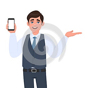 Trendy young businessman in vest suit showing blank mobile, cell phone and pointing to copy space. Person holding smartphone.