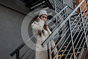 Trendy young cute woman in a fashionable knitted warm hat in a stylish milk-colored fur coat made of faux fur is standing in the