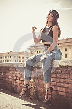 Trendy young girl sitting on a low wall outdoors. Jeans, sandals and hat, urban style. In the city on a brick wall