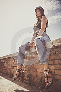 Trendy young girl sitting on a low wall outdoors. Jeans, sandals and hat, urban style. In the city on a brick wall