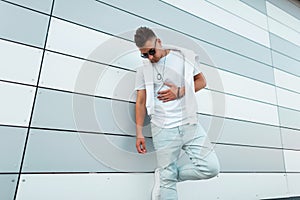 Trendy young hipster man in a fashionable white t-shirt in dark sunglasses in ripped jeans posing near a modern building on the